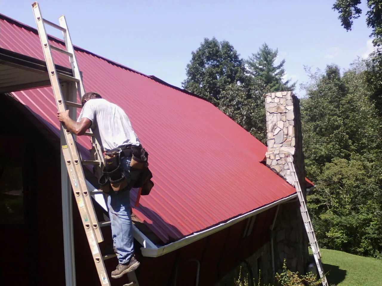 Roofer on a ladder beside a red metal roof and stone chimney — Soffit & Fascia Repair by Roofing Brothers in North Conway